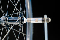 Close-up of a 700c HED Vanquish V45 Carbon Aero Wheelset axle with metal spokes and a black hub, showing the axle partially inserted into a metal frame dropout against a dark background. This wheelset features a 22.4 mm internal width and weighs just 1653g, making it both sturdy and lightweight for performance cycling.