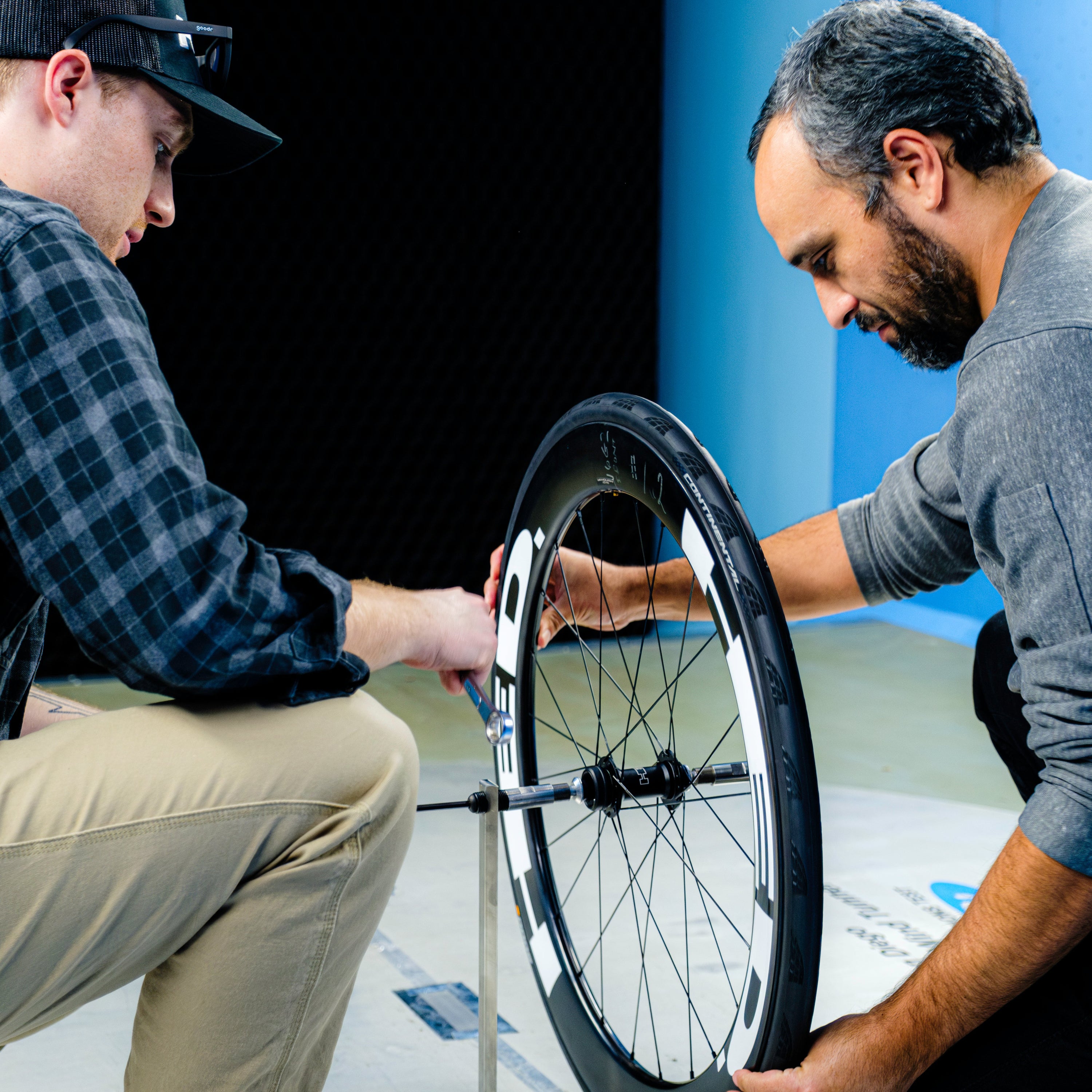 Two people kneel on the floor in a brightly lit indoor space with blue and black walls, using a ruler and tool to measure the alignment of a HED Vanquish V62 Carbon Aero Wheelset.