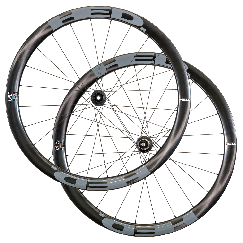 Two overlapping black and gray HED Vanquish V45 Carbon Aero wheels with thin spokes, visible hub mechanisms, and sleek aerodynamic rims are arranged against a white background, highlighting the advanced design of this premium carbon wheelset tailored for all-road riding.