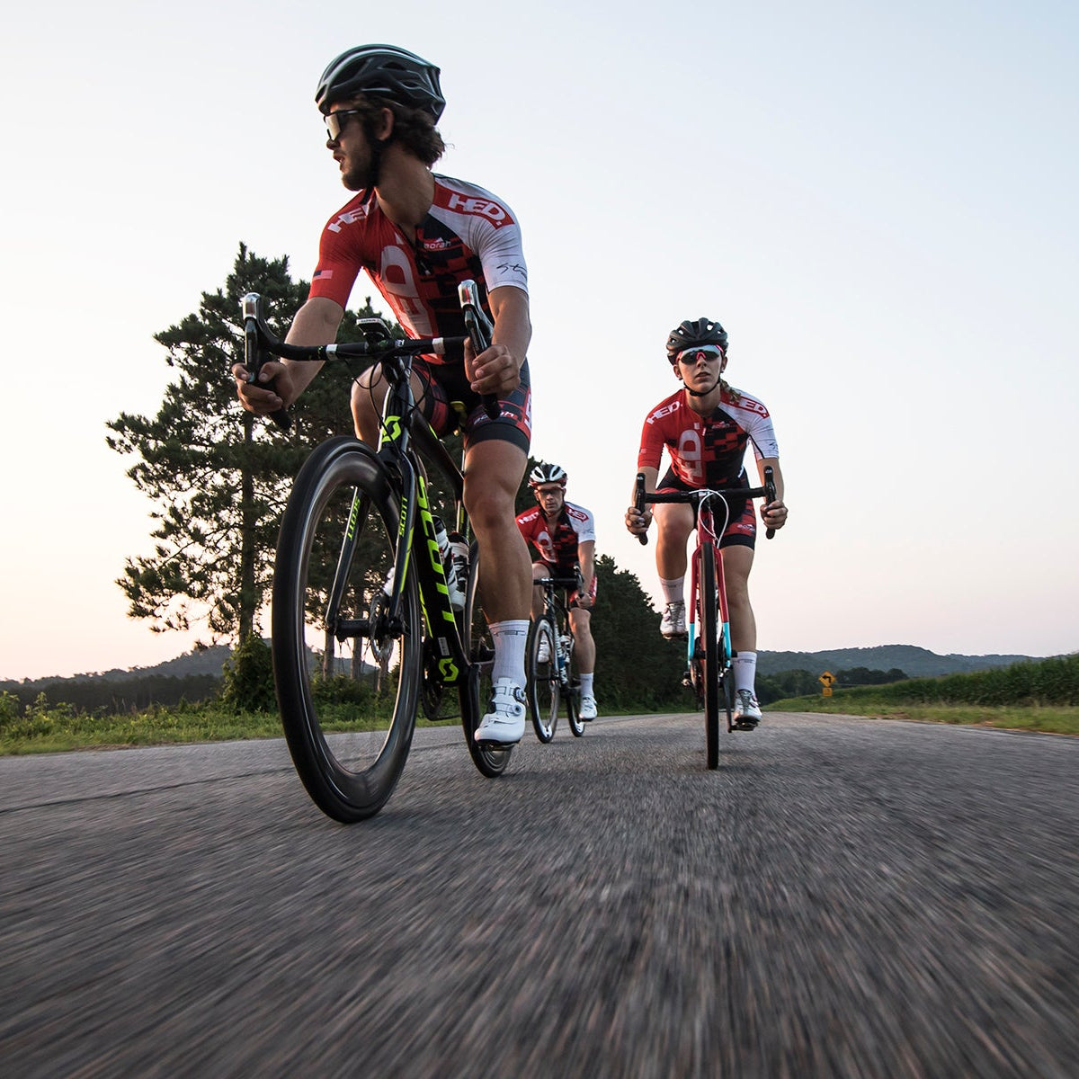 Four cyclists in matching red and white gear ride on a rural road at sunset, framed by trees and rolling hills. The low, close camera angle highlights the sleek HED Vanquish V62 Carbon Aero Wheelsets on their bikes as they push the pace—perhaps training for an upcoming triathlon.
